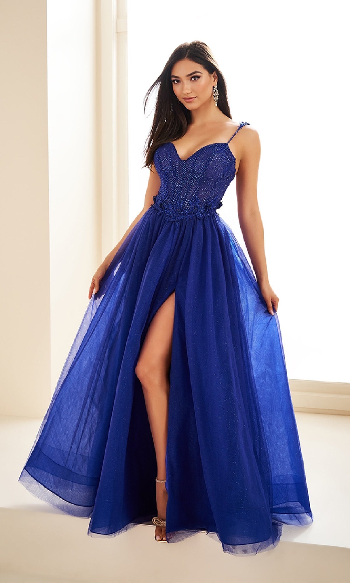 A woman stands indoors wearing the Ellie Wilde EW36058, a sleeveless floor-length royal blue prom dress with a beaded corset bodice and a high slit.